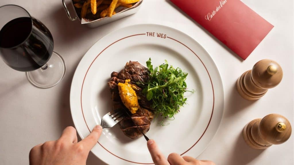 A gourmet steak dish with greens on a white plate at The Wes restaurant, with a glass of red wine and condiments nearby.