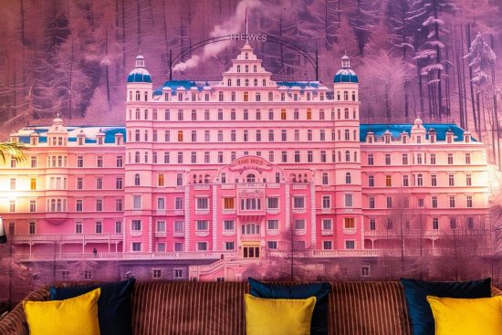 Pink hotel with a castle-like design and multiple towers, set against a forest background, with warm lighting and modern furniture in the foreground.