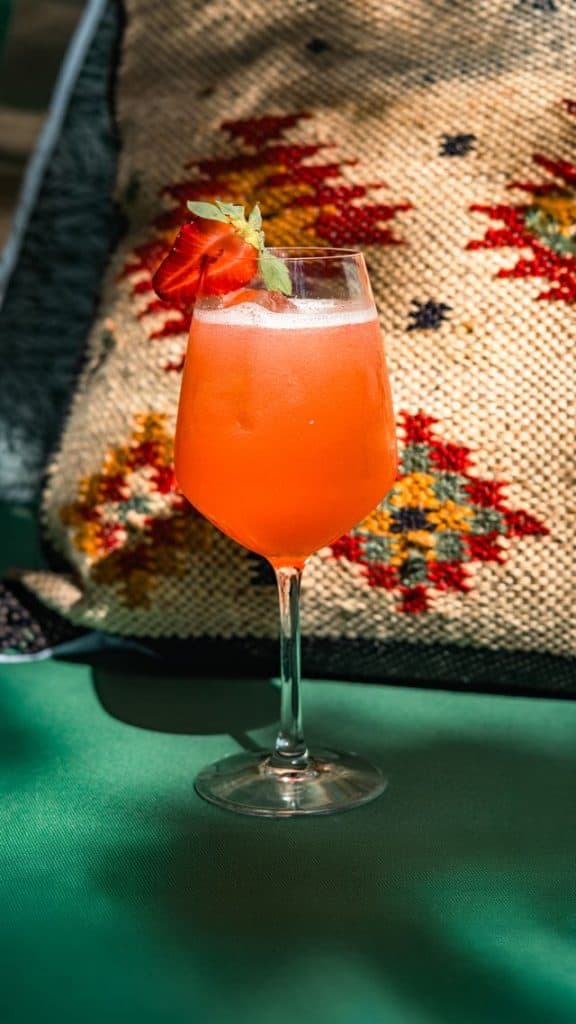 A vibrant pink cocktail garnished with a slice of orange and a mint leaf, served in a tall glass on a green table.