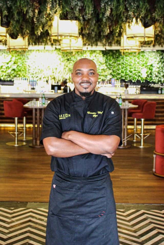Smiling chef in black uniform standing confidently in a modern restaurant with lush green decor and warm lighting.