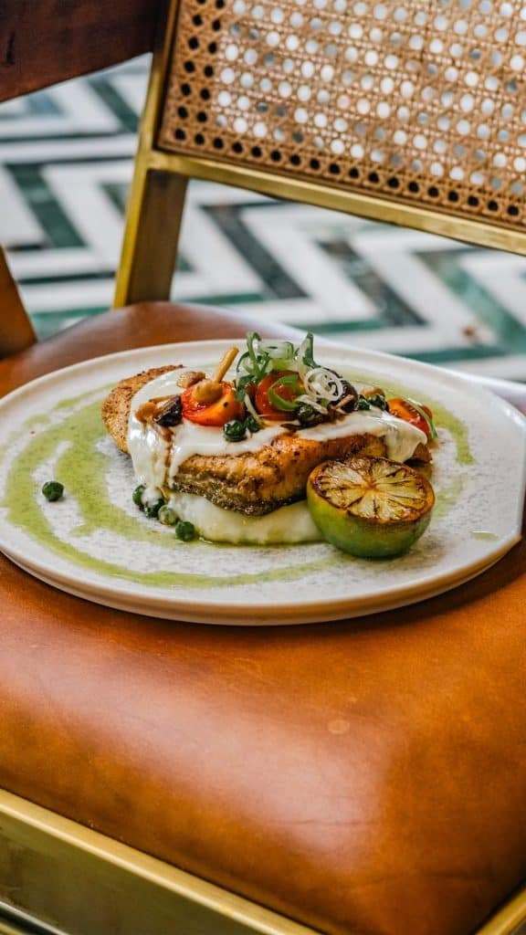 Delicious gourmet fish dish served with lemon, herbs, and sauce on a white plate, with a stylish chair in the background.