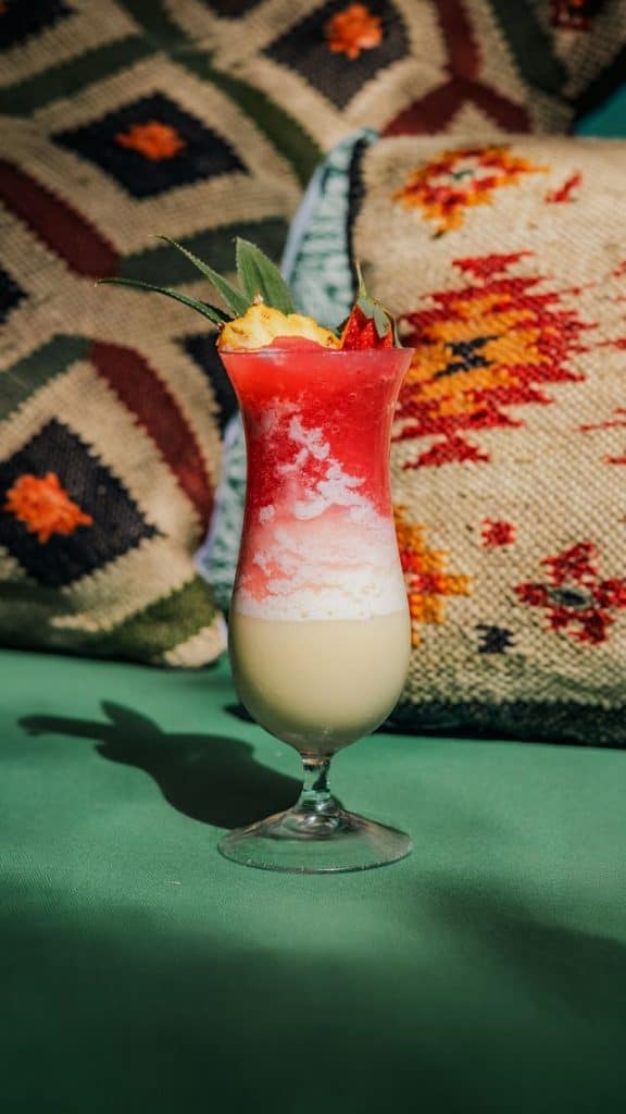 Vibrant layered cocktail with red, white, and pink hues garnished with a lime wedge, set against a patterned textile background.