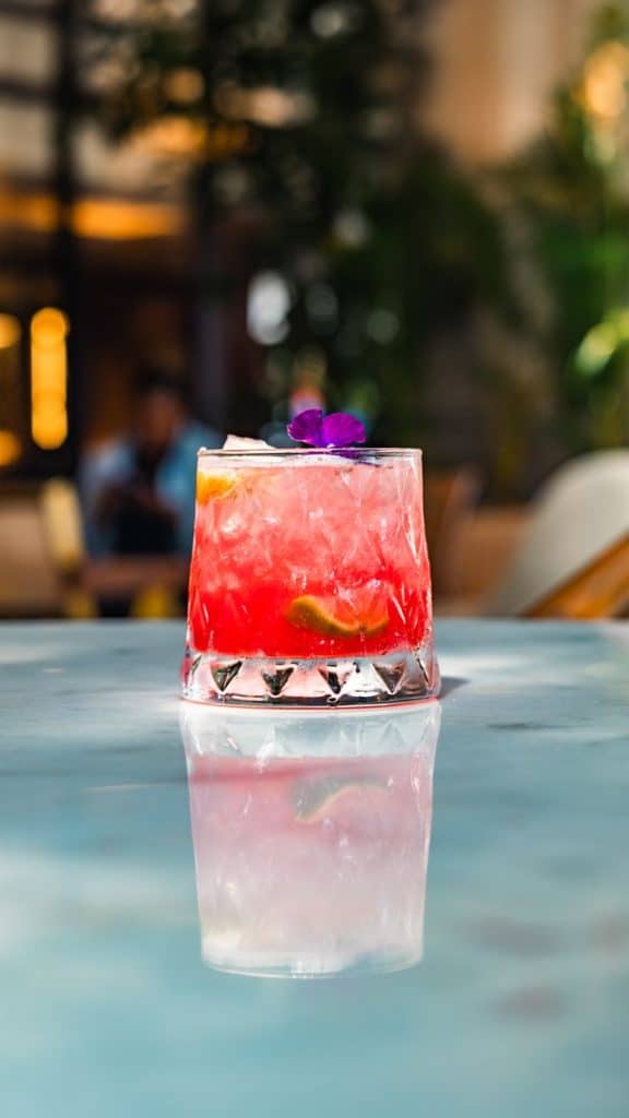 A vibrant pink cocktail garnished with a purple flower and lime slice, served in a clear glass on a reflective surface with a blurred outdoor background.