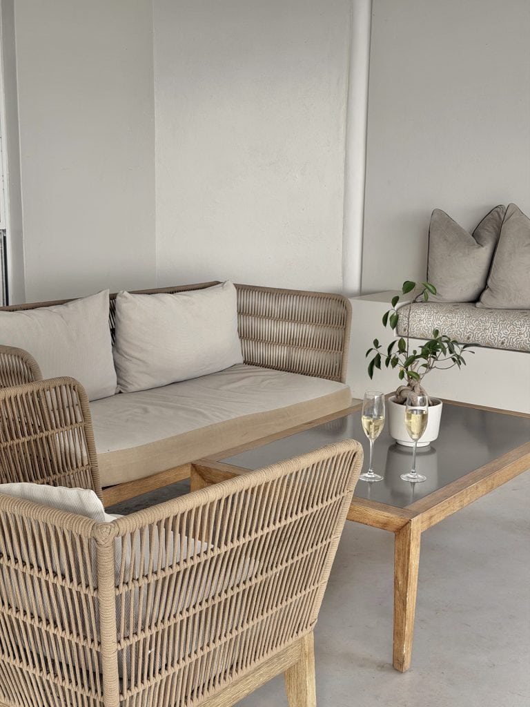 Modern living room with beige rattan sofa, grey cushions, and a wooden coffee table with two glasses of white wine and a potted plant.
