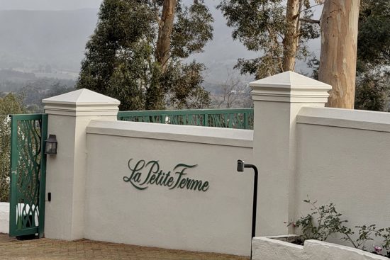 Entrance gate and wall with 'La Petite Ferme' sign, surrounded by trees and misty background.