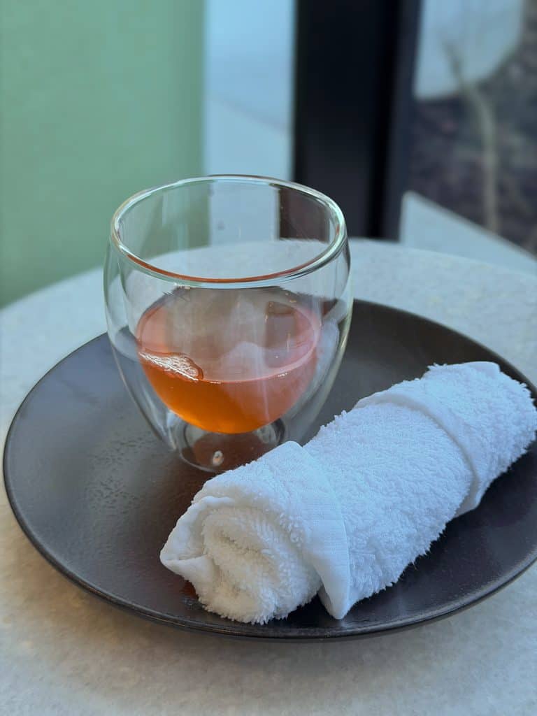 Glass of rosé wine with a white towel on a black plate near a window, offering a relaxing spa vibe.