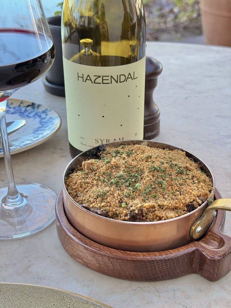 A bottle of Hazendal wine, a glass of red wine, and a baked dish on a table at Hazendal Wine Estate in Stellenbosch.