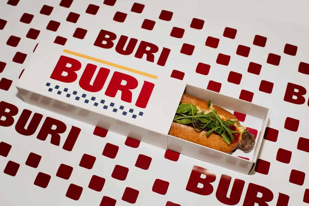 Buri sandwich in a branded box with a South African theme, featuring fresh greens and a bun, on a patterned tablecloth.