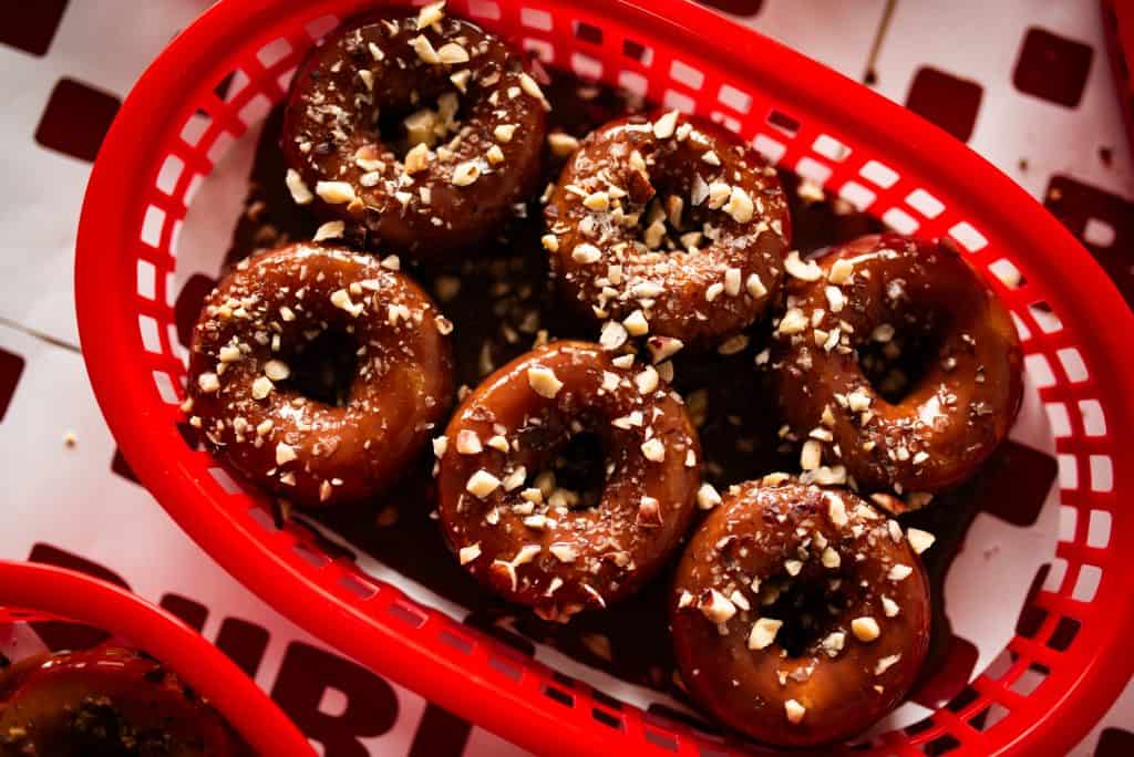A close-up image of glazed donuts topped with chopped nuts, arranged in a red basket. The donuts are shiny and appear freshly baked, highlighting their sweet and crunchy texture.